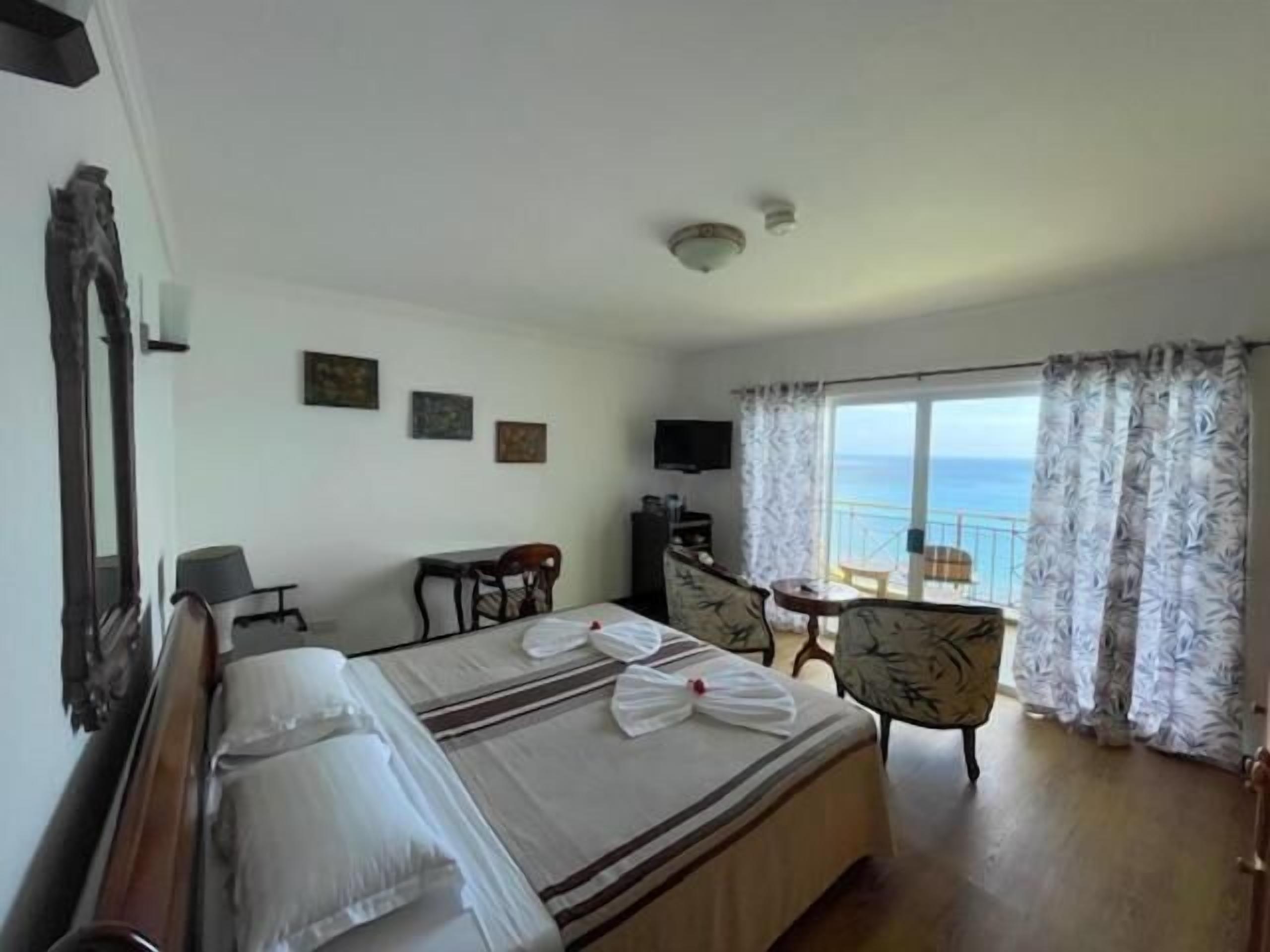 A hotel room with a bed, a desk, a chair, a TV, and a balcony with a view of the sea.