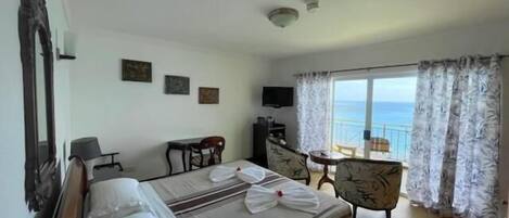 A hotel room with a bed, a desk, a chair, a TV, and a balcony with a view of the sea.