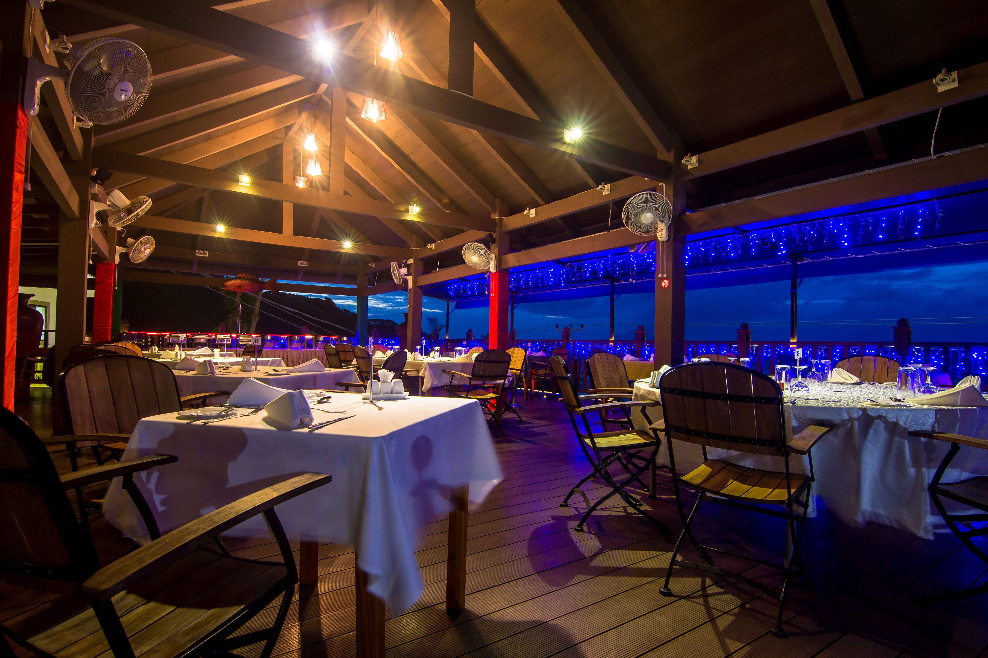 Un espacio exterior para comer con mesas dispuestas para una comida, vigas de madera en el techo y luces colgantes.