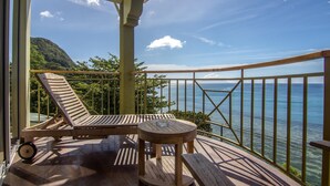 A balcony with a wooden table and two lounge chairs, overlooking a clear blue sea and a green hillside. - Treasure Cove (Mahé Island)