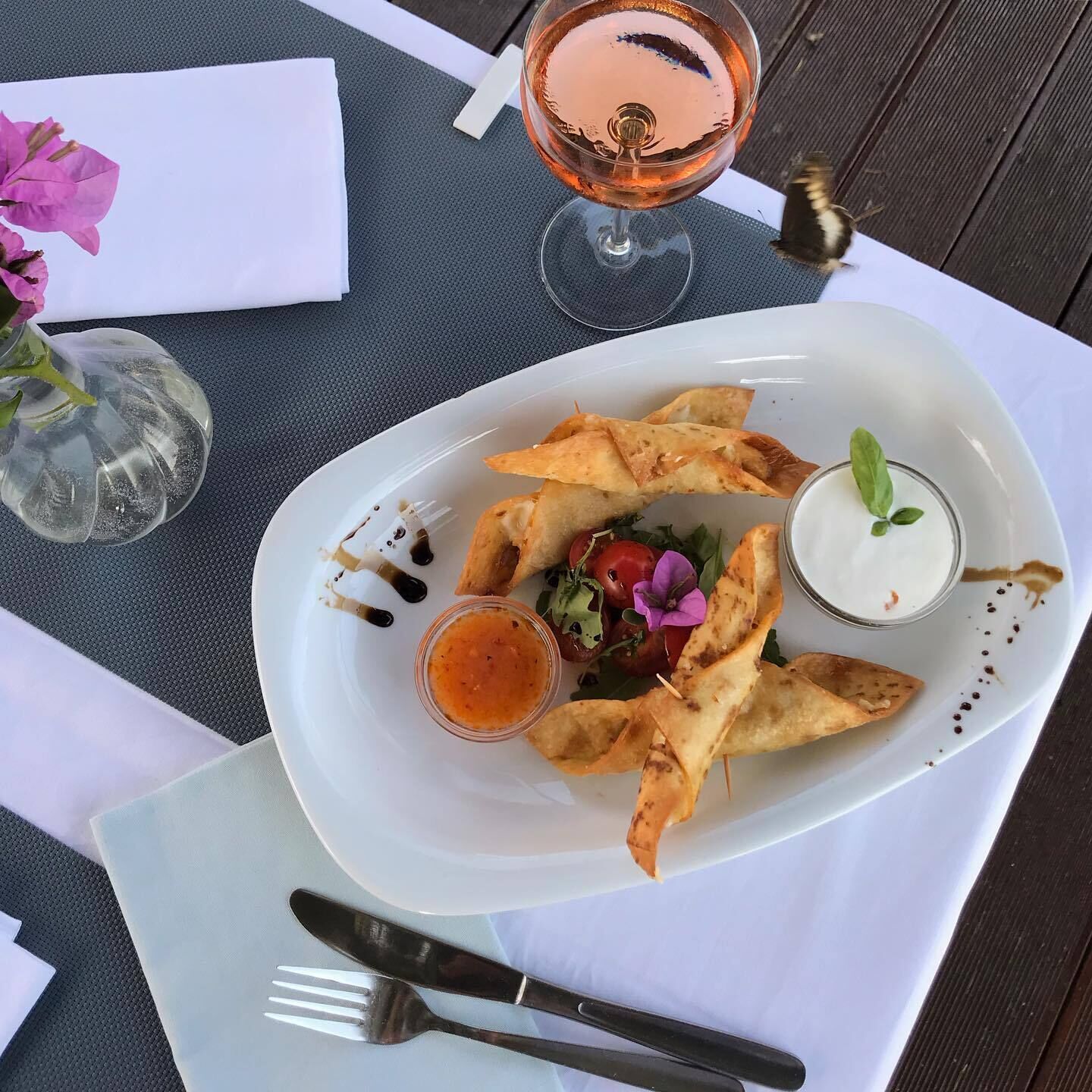 A white plate with food, a glass of drink, and a vase with flowers on a table.