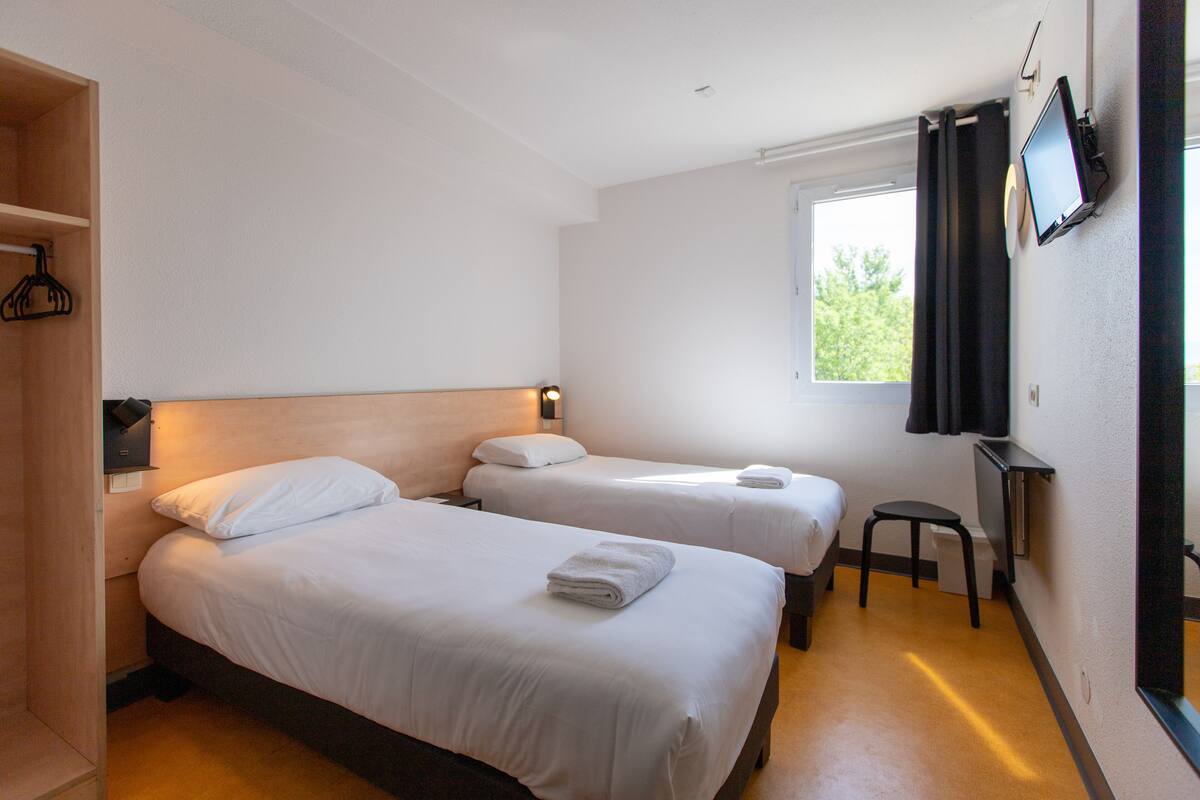 A hotel room with two beds, a TV mounted on the wall, a small stool, and a window with curtains.
