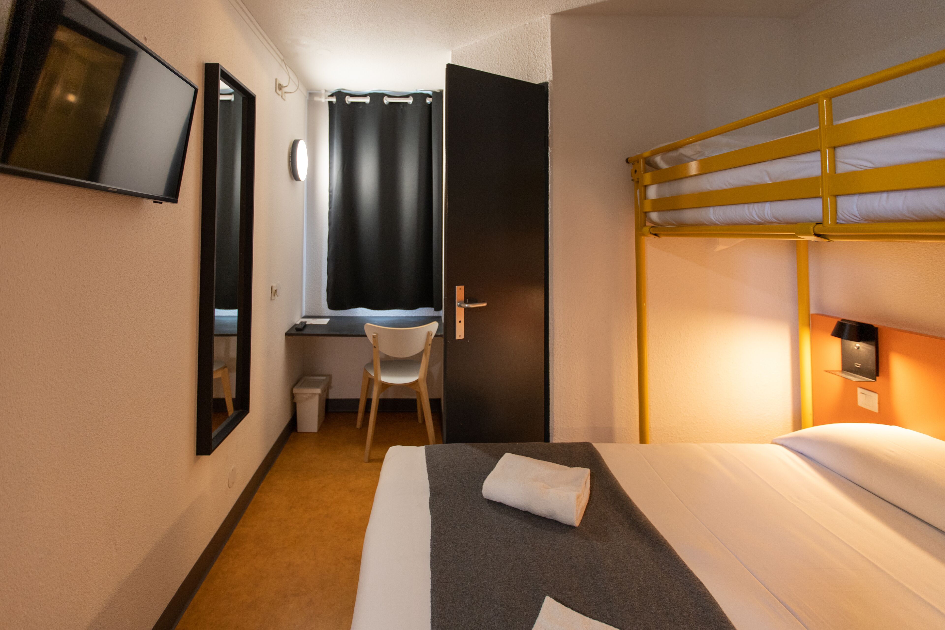 Une chambre d’hôtel avec un lit superposé, un bureau avec une chaise, une télévision et une fenêtre avec des rideaux.