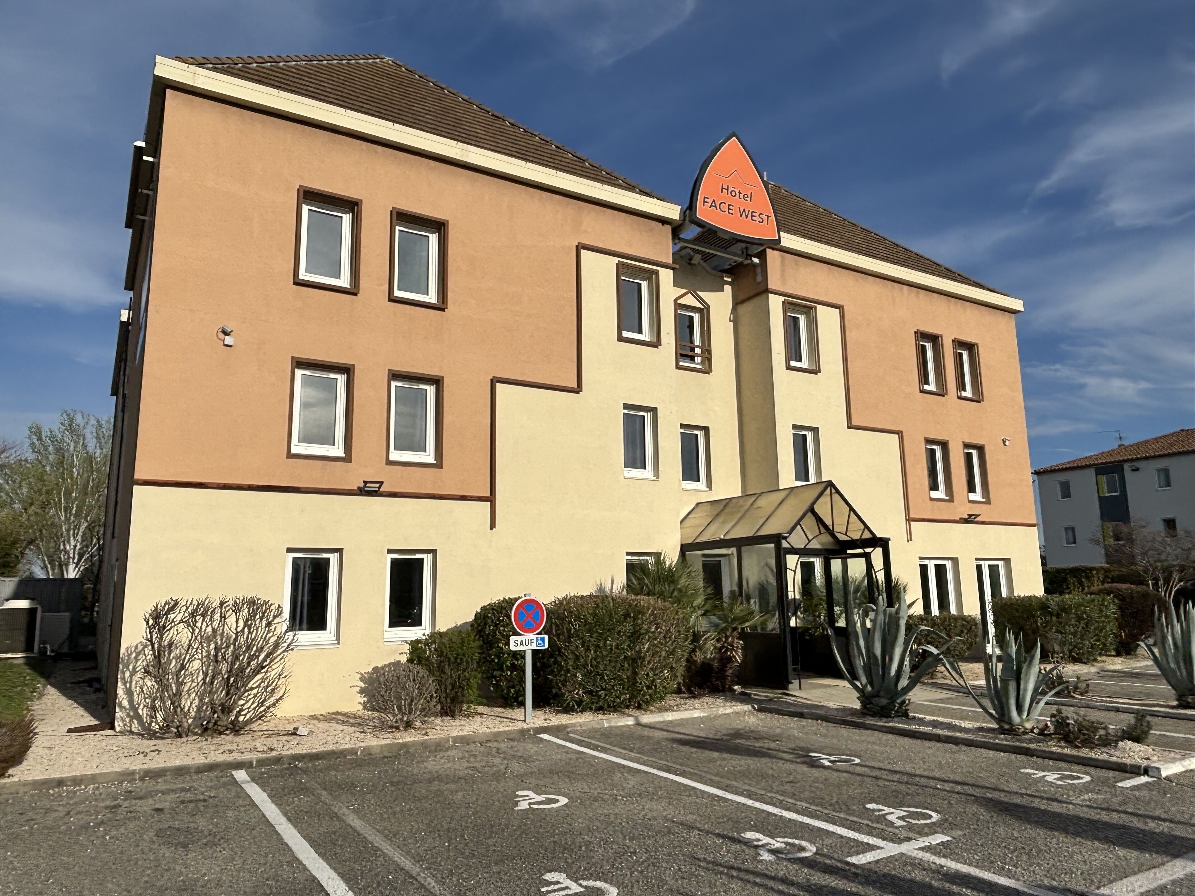 Edificio de hotel con un letrero que dice "Hotel FACE WEST". Presenta un diseño moderno con múltiples ventanas y un estacionamiento con espacios delimitados.