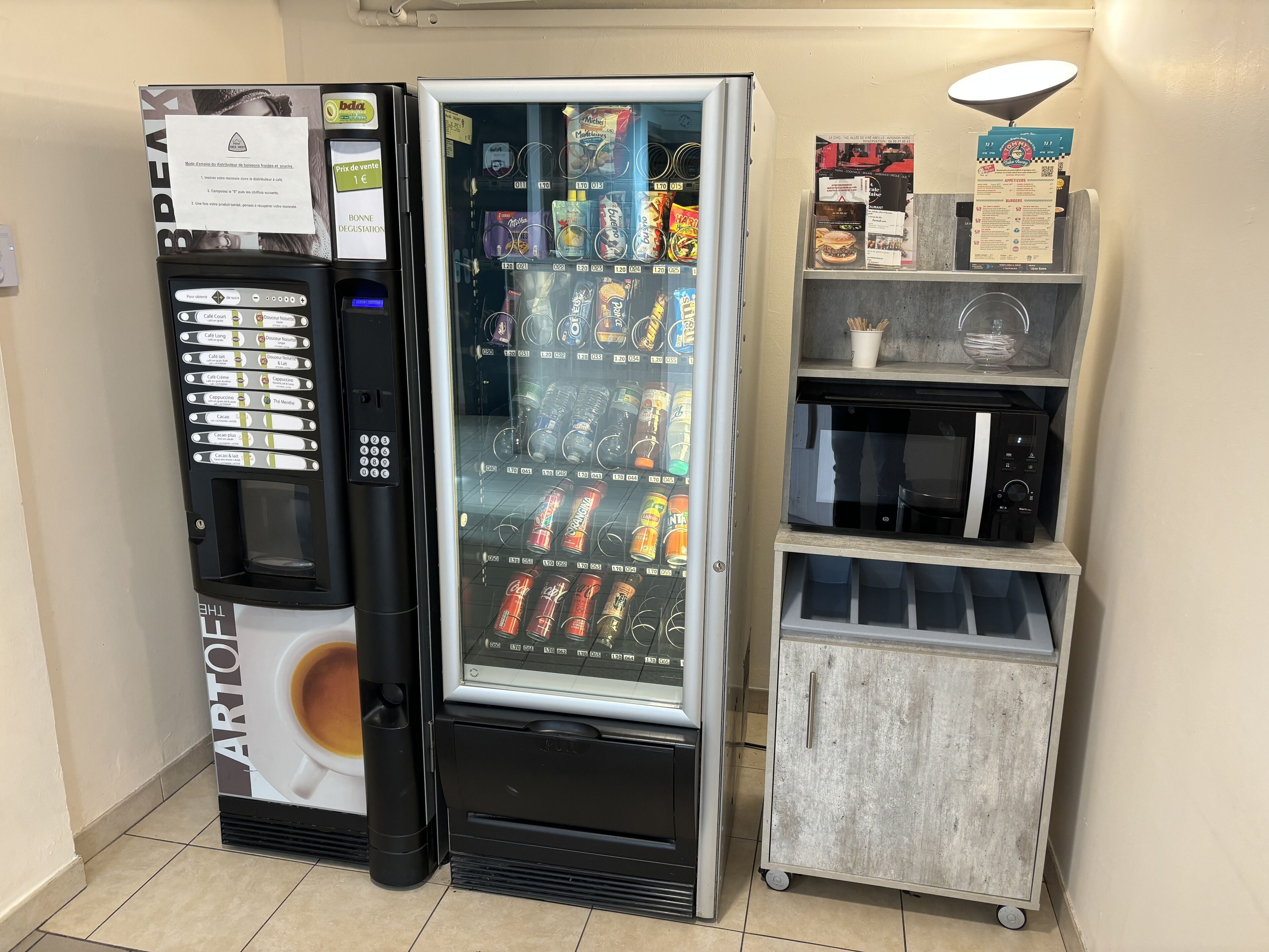 A vending machine with a beverage dispenser and a refrigerated section, next to a small cart with a microwave and a shelf with condiments.