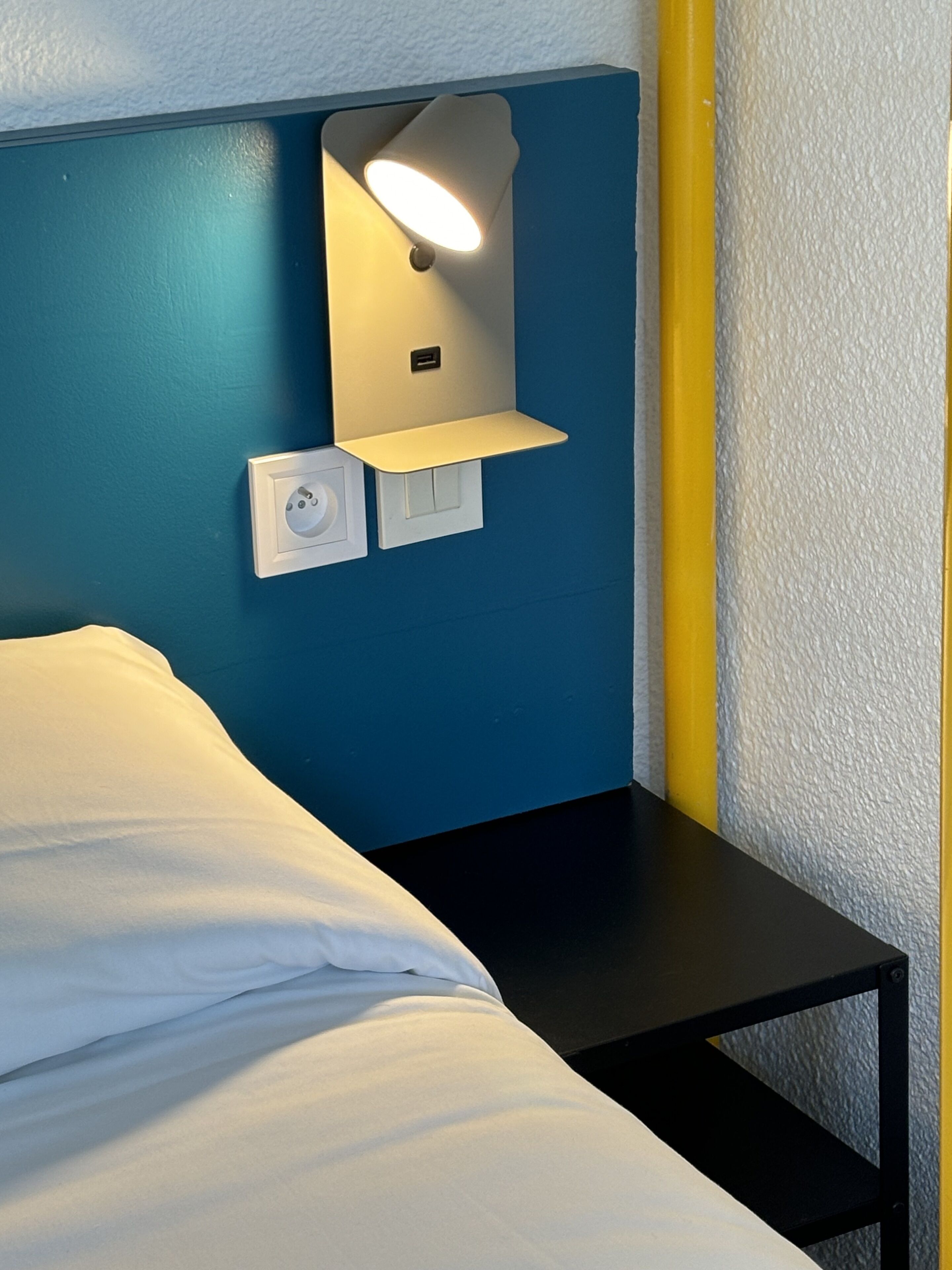A bed with white linens, a nightstand, a wall-mounted lamp, and a power outlet.
