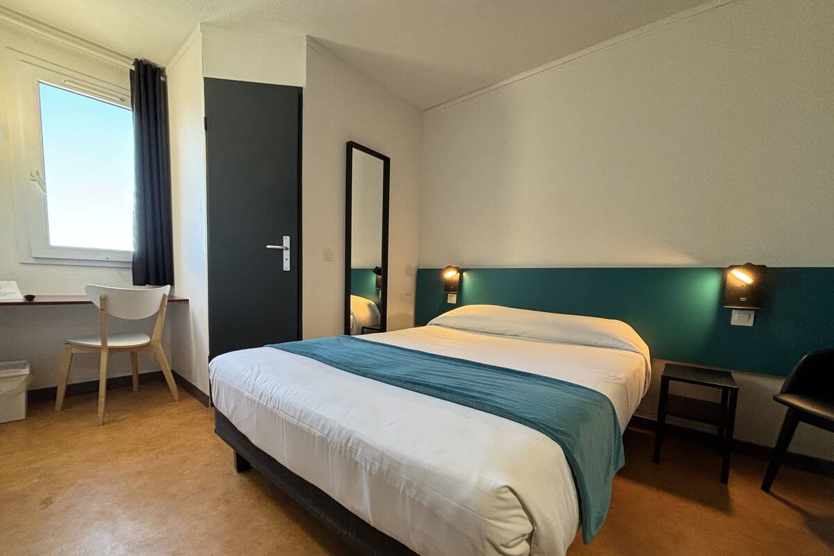 A hotel room with a bed, a desk, a chair, a mirror, and a window with curtains.