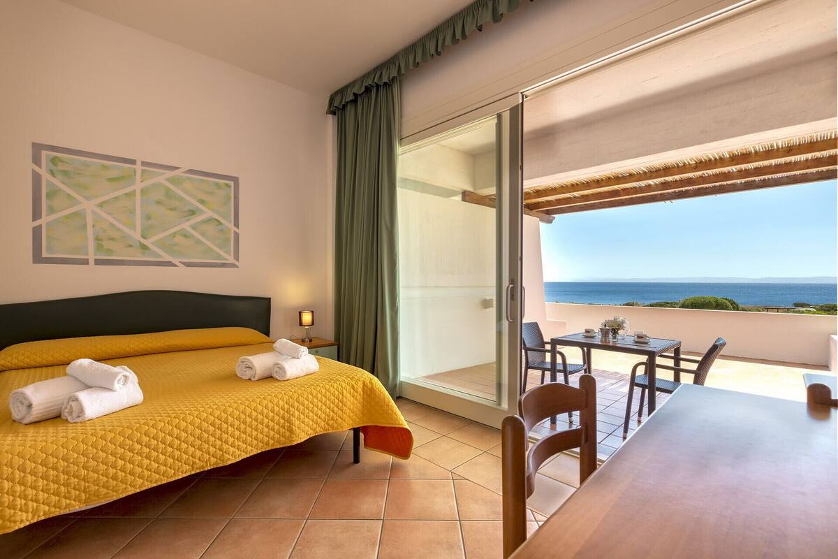 A hotel room with a bed, a table, chairs, a view of the sea, and a painting on the wall.