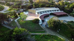 Vista aerea di un edificio moderno con tetto curvo, circondato da vegetazione e un'area pavimentata con posti a sedere.