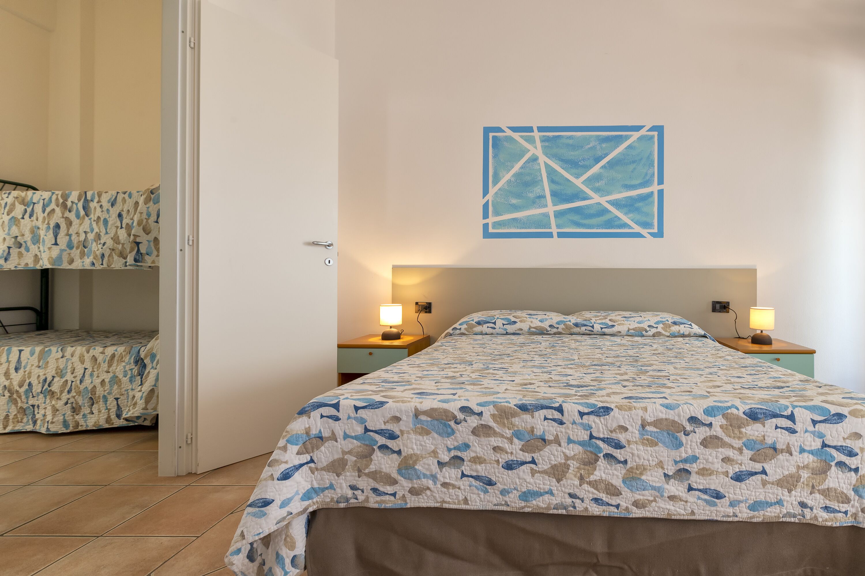 A bedroom with a bed, two bedside tables with lamps, a wall art piece, and a door leading to another room.