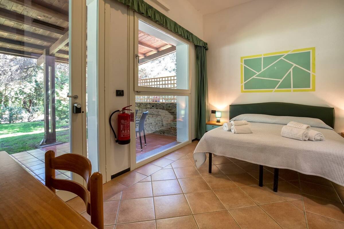 A bedroom with a bed, a chair, a table, a fire extinguisher, and a view of the outdoors.