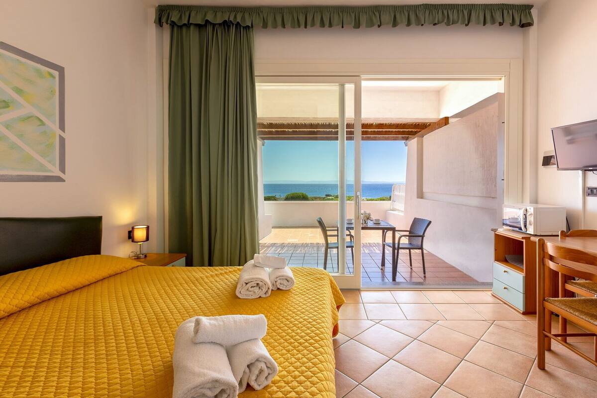 A hotel room with a bed, a desk, a chair, a TV, and a balcony with a view of the sea.