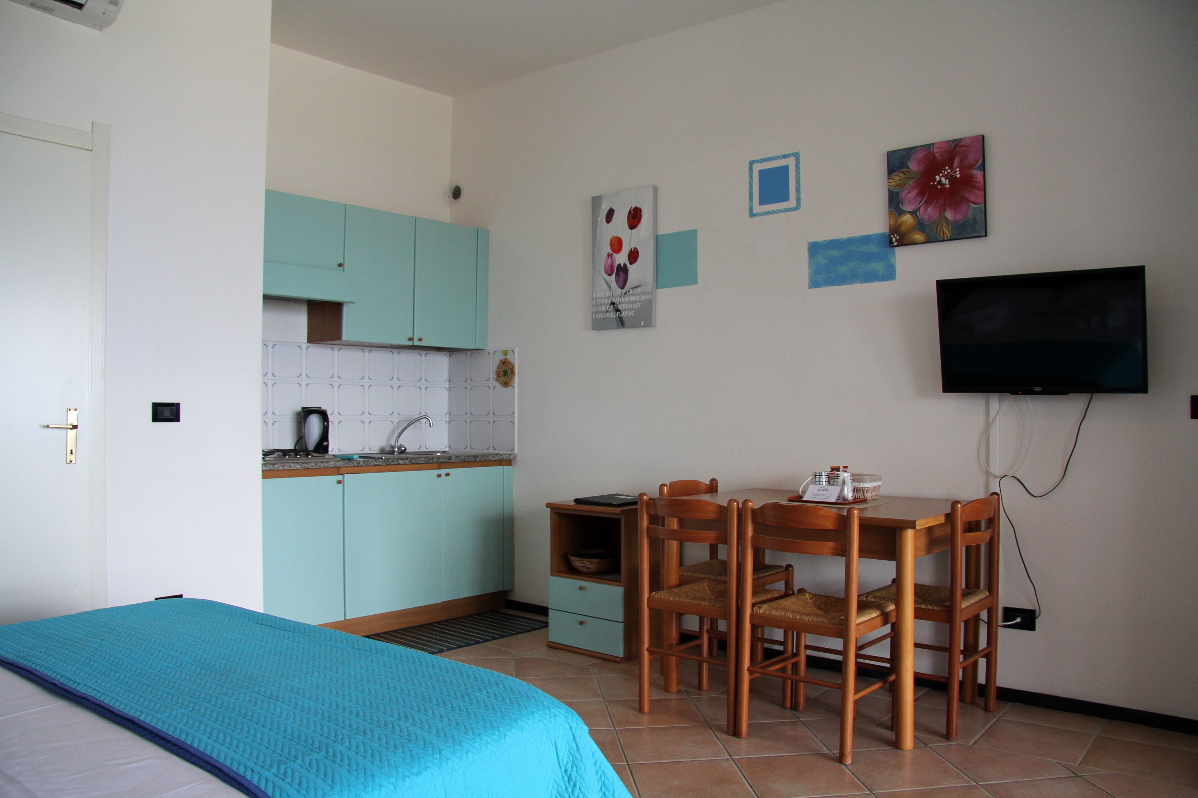 A bedroom with a bed, a kitchenette with teal cabinets, a dining area with a table and chairs, and a TV mounted on the wall.