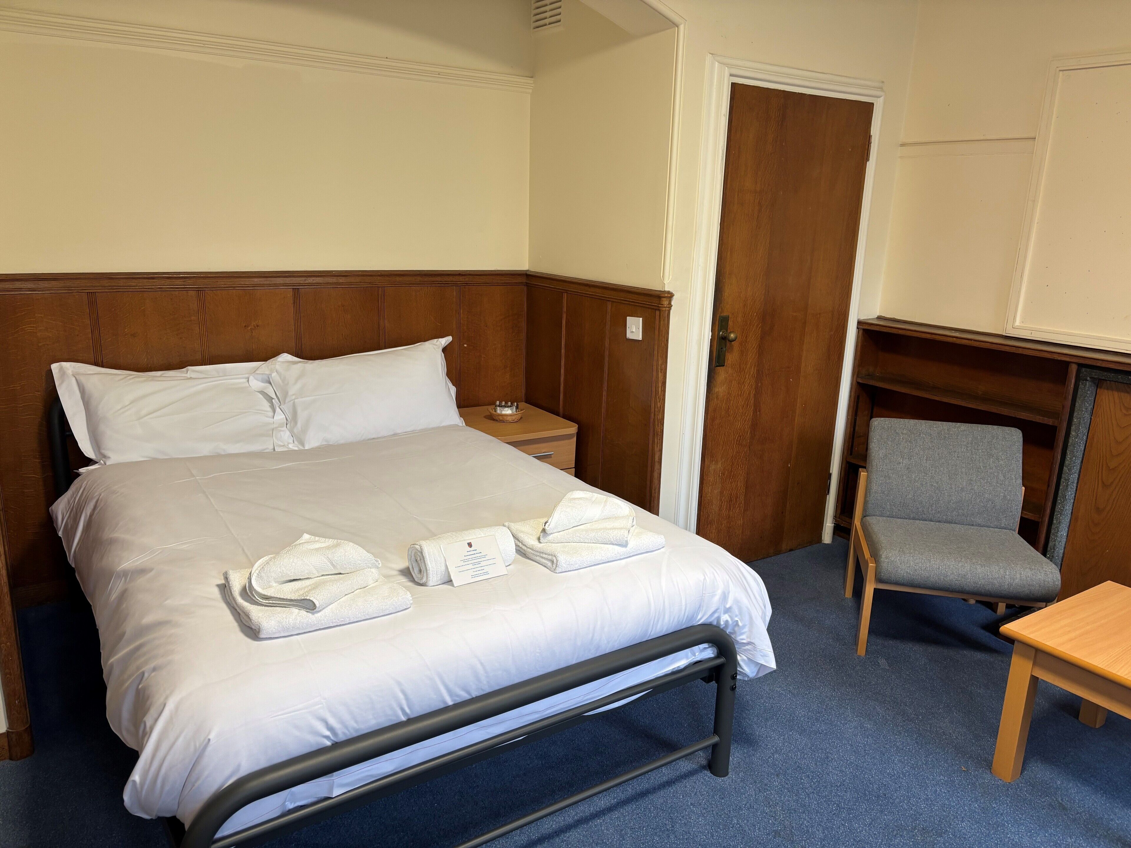 A hotel room with a bed, a chair, a small table, and a wooden wardrobe.