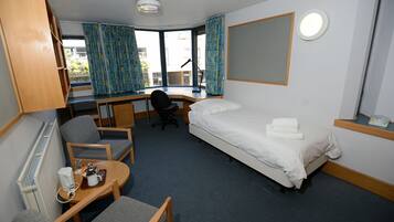 Una habitación de hotel con una cama, un escritorio, una silla, una mesita con una taza y un teléfono, y una ventana con cortinas.