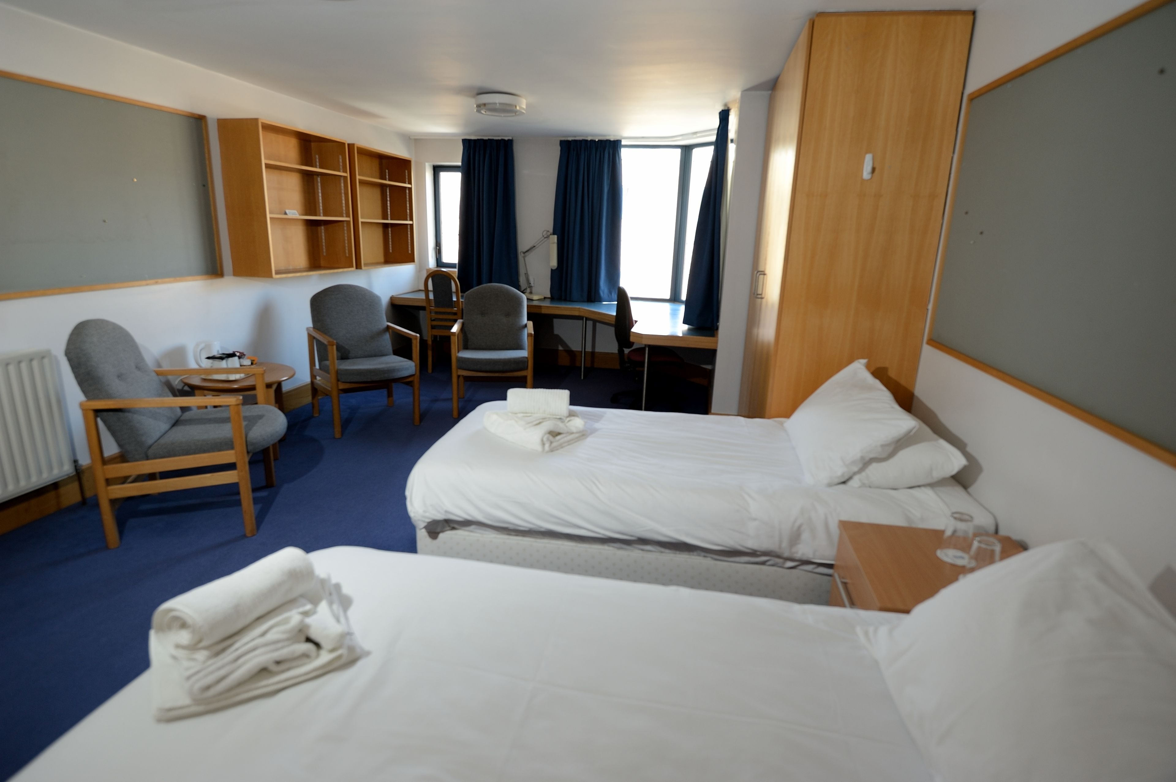 A hotel room with two beds, a desk, a chair, and a bookshelf.