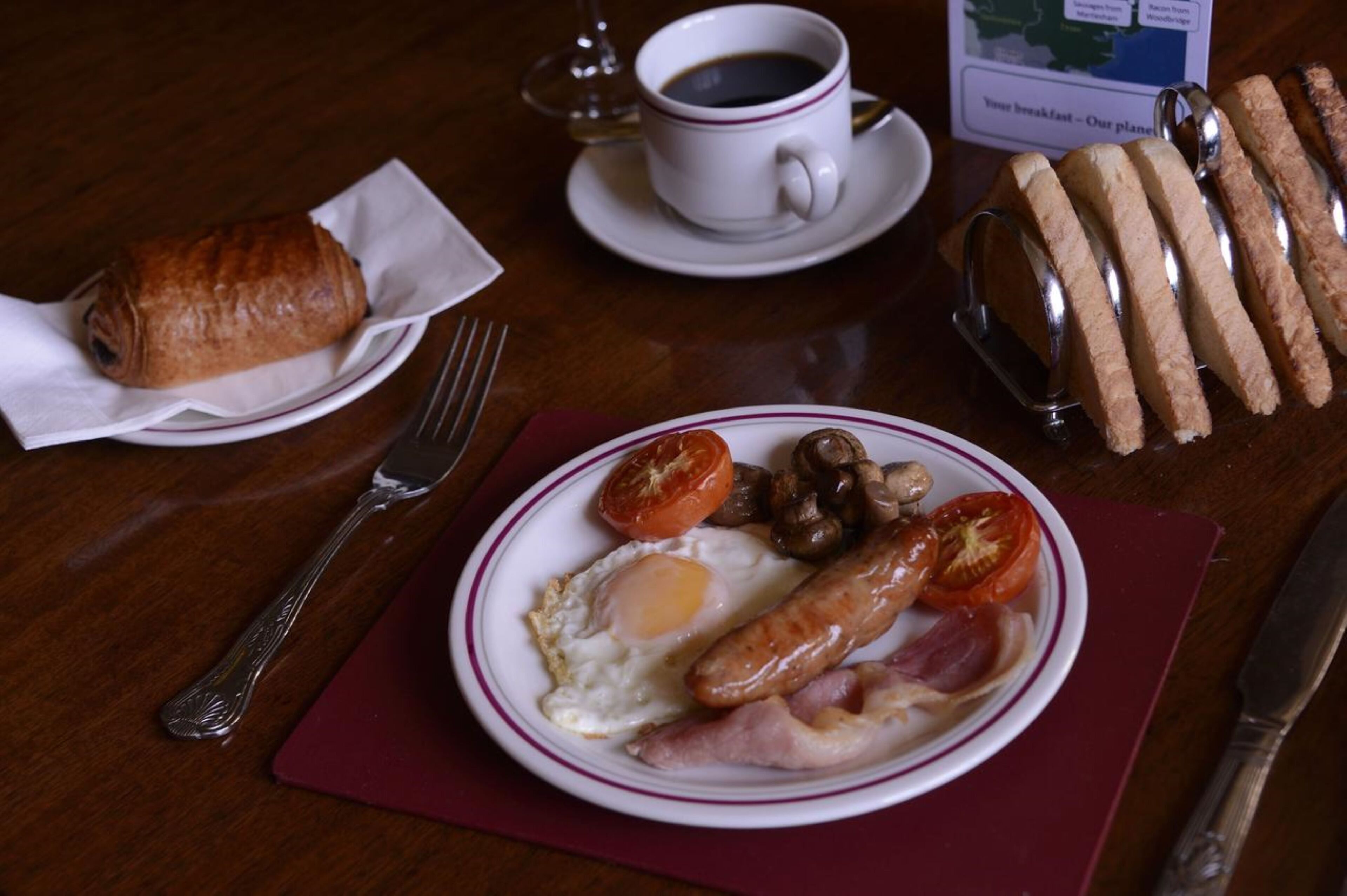 Un plat amb ous, botifarreta, tomàquets i bolets, un croissant i una tassa de cafè damunt d'una taula de fusta.