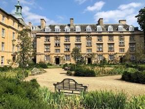 Stately building with a central entrance and two wings, set amidst serene grounds with a gravel path and a bench for relaxation.