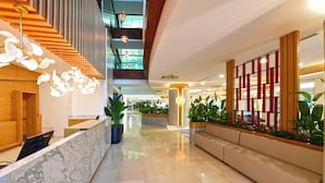 Un moderno vestíbulo de hotel con una recepción de mármol, detalles de madera, un área de descanso y una escalera que conduce a un nivel superior.