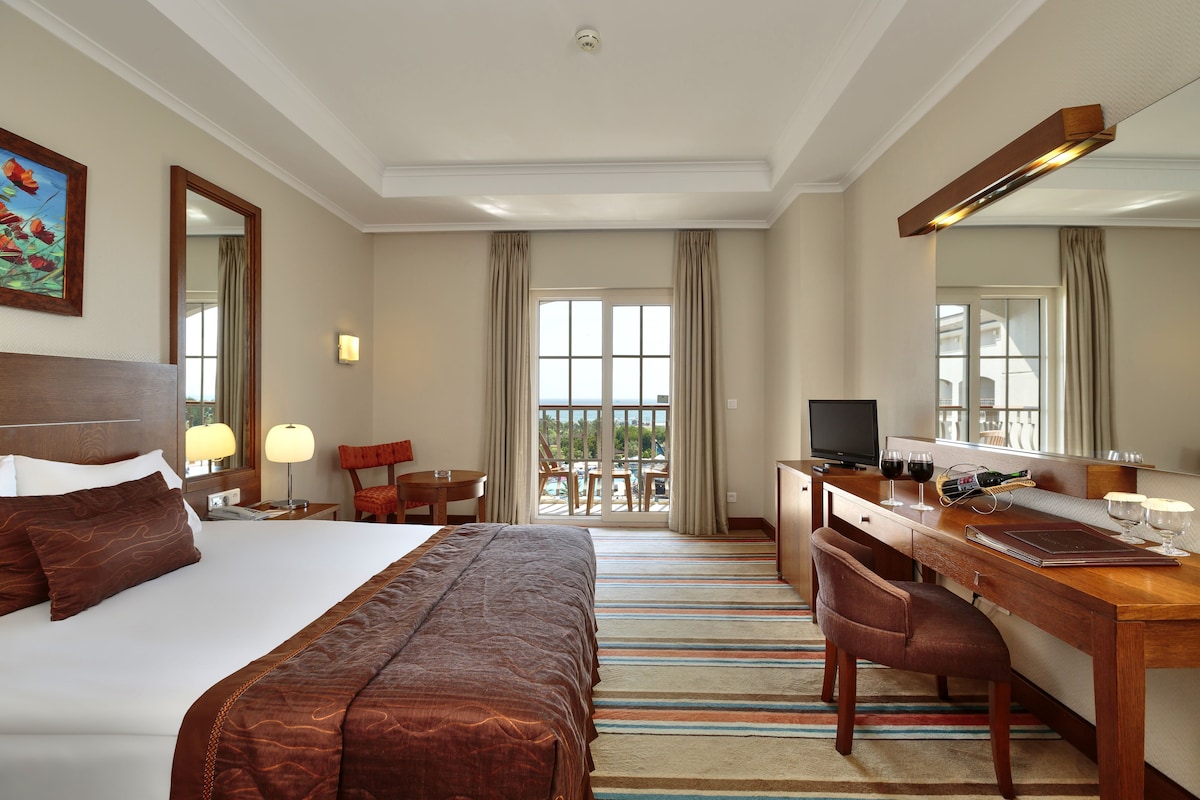 A hotel room with a large bed, a desk, a chair, a television, and a balcony with a view.