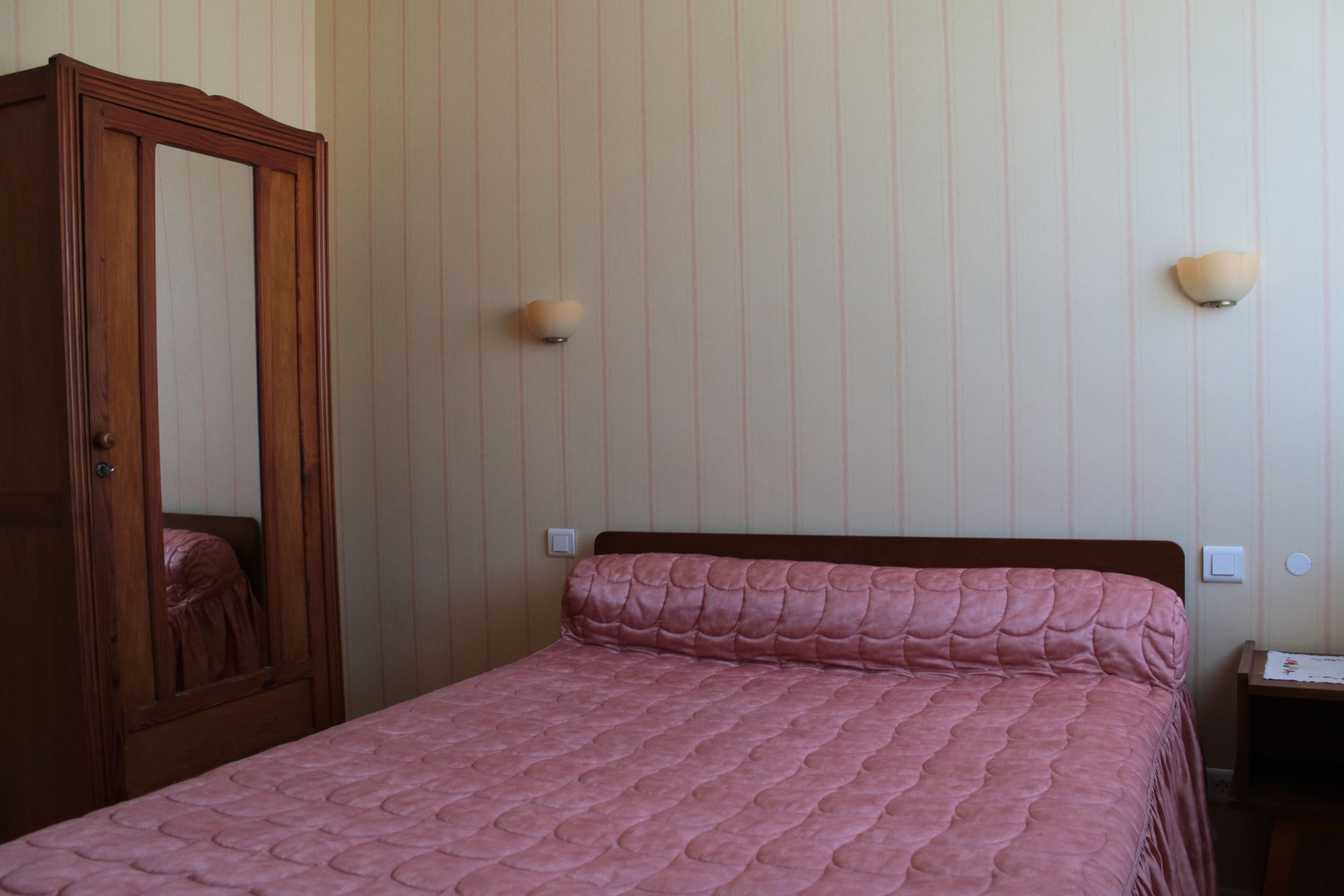 A bedroom with a wooden wardrobe, a pink quilted bedspread, and two wall-mounted light fixtures.