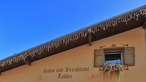 A building with a sign reading "Room and Breakfast Ladin." It features a brick facade, wooden shutters, and hanging flower baskets. - Garnì Ladin (San Giovanni di Fassa)