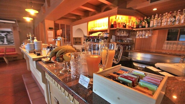 A bar area with a counter displaying various items like a pitcher of orange juice, a banana, and a tray of colorful pens. - Garnì Ladin (San Giovanni di Fassa)