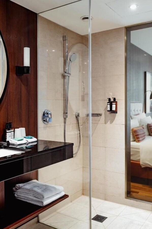 Une salle de bain moderne avec une douche en verre, un meuble-lavabo en bois foncé et un lit visible dans la pièce adjacente, agrémenté d’oreillers.