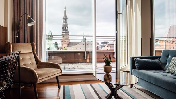 A modern living room with a sofa, armchair, and a glass coffee table. A large window offers a view of a cityscape with a prominent tower. - AMERON Hamburg Hotel Speicherstadt (Hamburg)