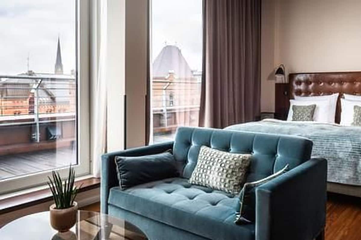 A hotel room with a large bed, a blue sofa, a wooden headboard, and a view of a cityscape through large windows.