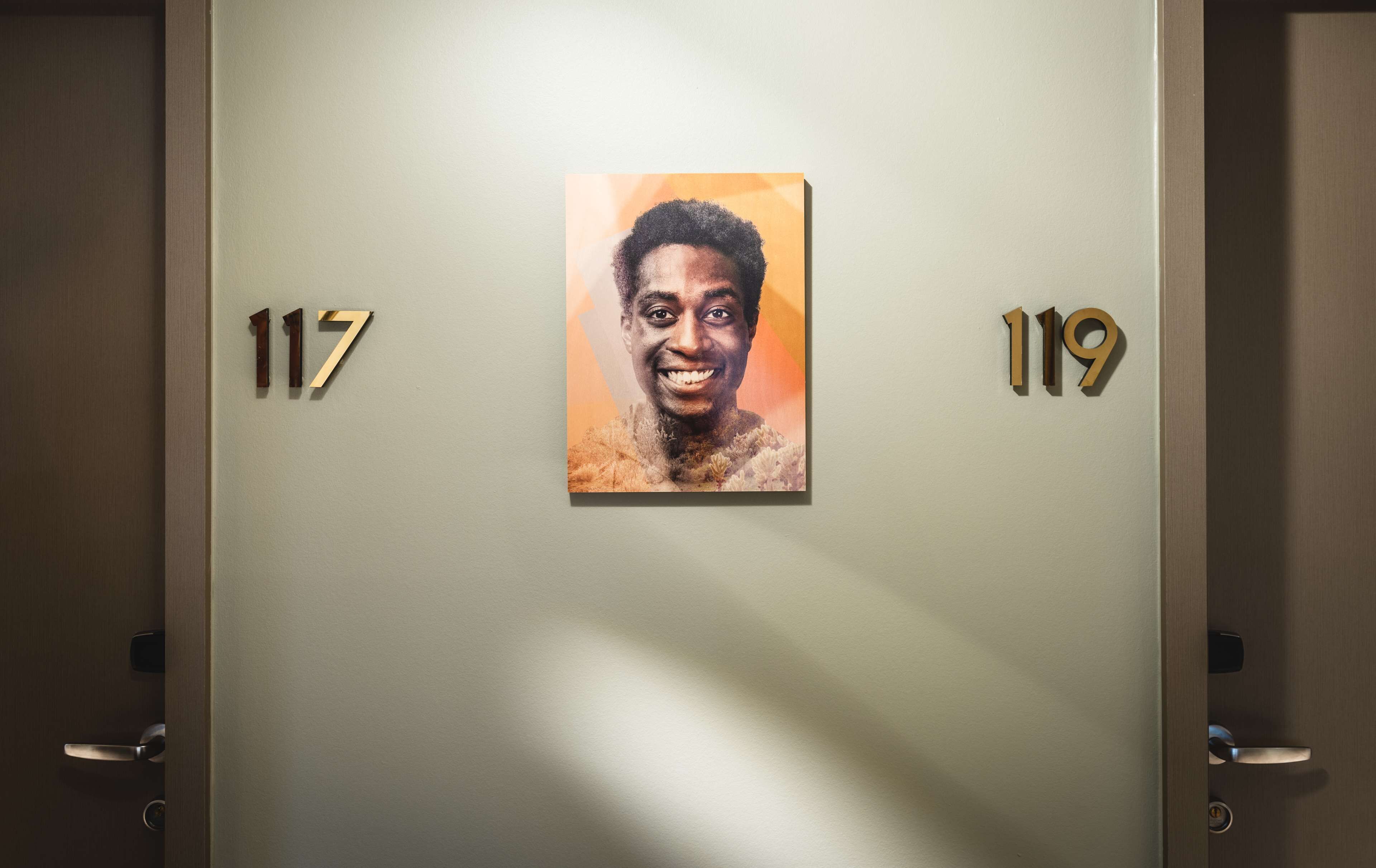 Un couloir avec des portes numérotées 117 et 119, orné d’un portrait encadré accroché au mur.