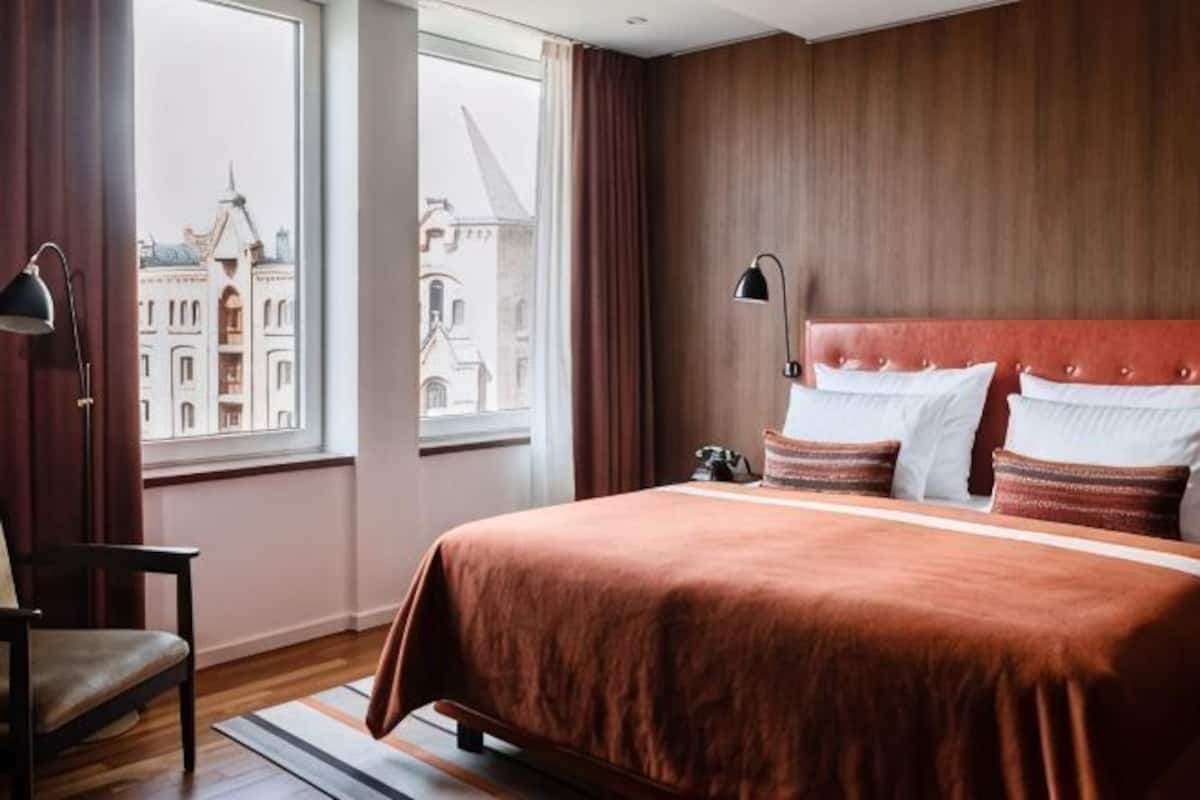 Ein Hotelzimmer mit einem großen Bett, einem Sessel, einem Nachttisch und Blick auf Gebäude durch ein Fenster.