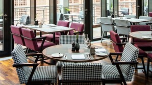 A restaurant with high ceilings, large windows, and a view of buildings. Tables are set with red and black patterned chairs. - AMERON Hamburg Hotel Speicherstadt (Hamburg)