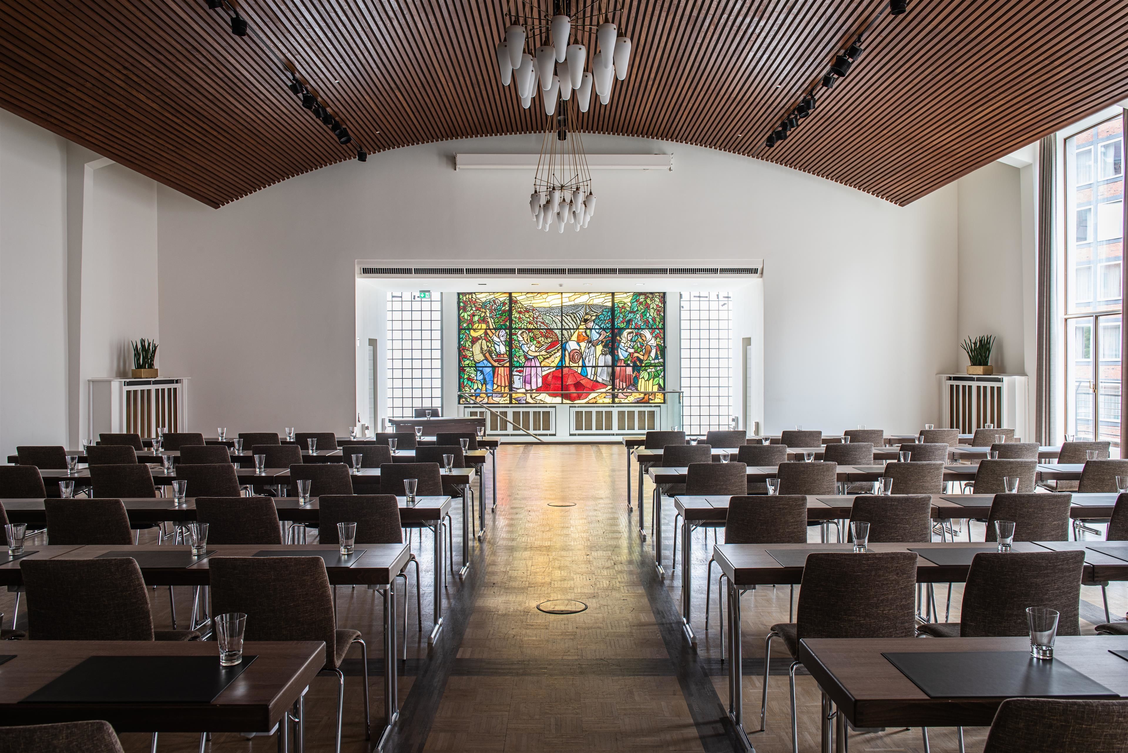 Une salle de conférence avec des rangées de tables et de chaises, une grande fenêtre à vitraux à l’avant et un plafond en bois orné de luminaires suspendus.