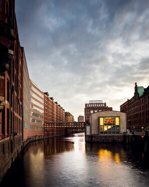 A canal with buildings on either side, a bridge, and a modern building with large windows reflecting light. - AMERON Hamburg Hotel Speicherstadt (Hamburg)
