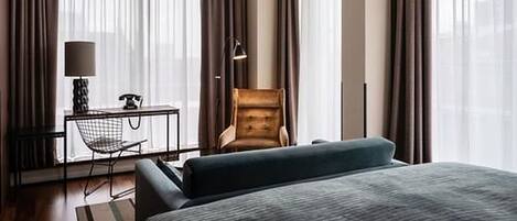 A hotel room with a bed, a desk with a lamp, a chair, and large windows with curtains.