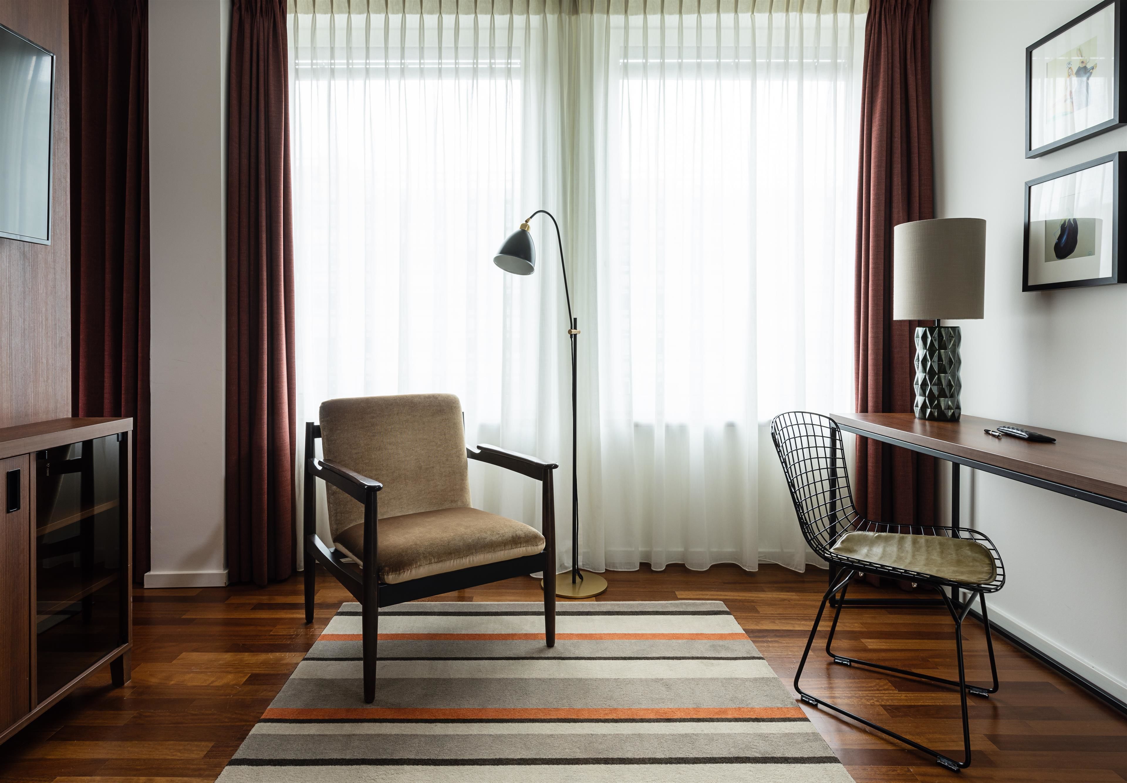 A room with a wooden floor, a sofa, a chair, a desk, a lamp, and a TV on the wall.