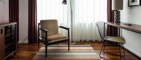 A room with a wooden floor, a sofa, a chair, a desk, a lamp, and a TV on the wall.