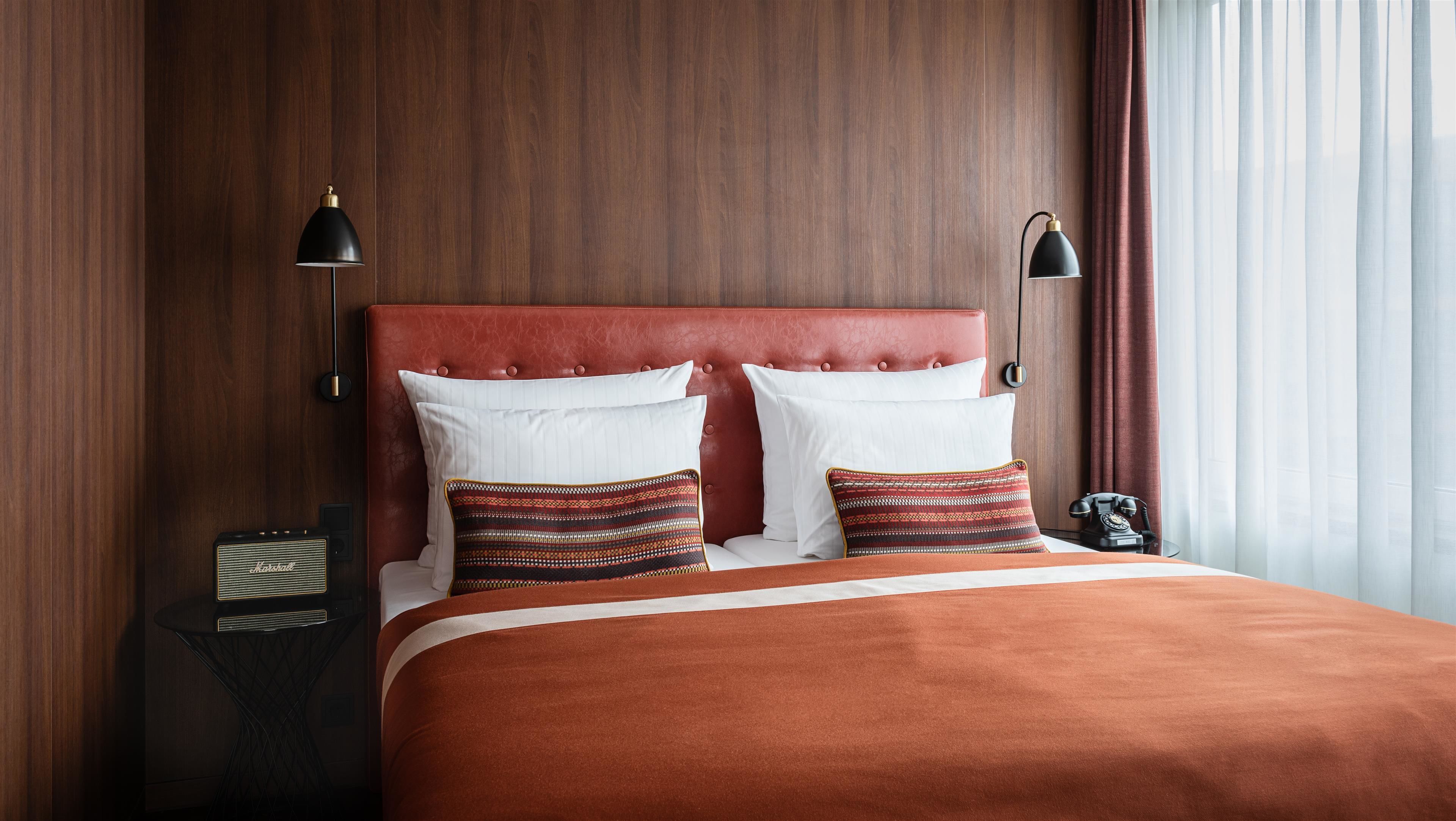 Une chambre d’hôtel avec une tête de lit en bois, deux oreillers blancs, un coussin à motifs, un plaid rouge, une lampe noire et un téléphone d’époque posé sur une table de chevet.