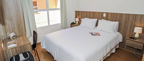 Habitación de hotel con una cama grande, cabecera de madera, dos mesitas de noche con lámparas, un escritorio con silla y una ventana con cortinas.