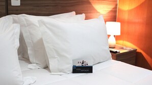 A neatly made bed with white linens, a bedside lamp, and a small card on the nightstand. - The Hill Executive (São Carlos)