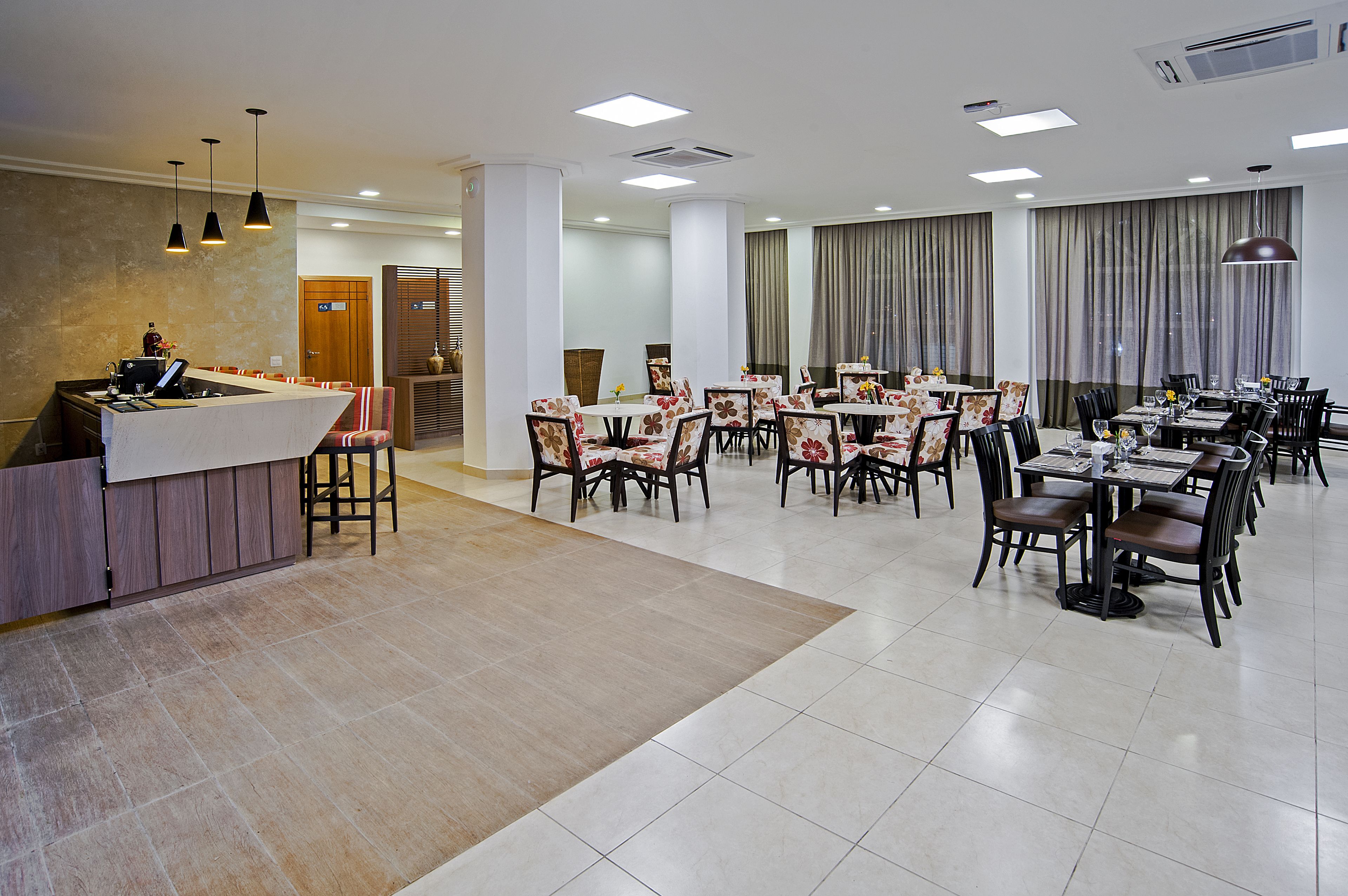Um amplo espaço de refeições de hotel, com várias mesas e cadeiras, um balcão de recepção e uma grande janela com cortinas.