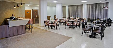 Um amplo espaço de refeições de hotel, com várias mesas e cadeiras, um balcão de recepção e uma grande janela com cortinas.
