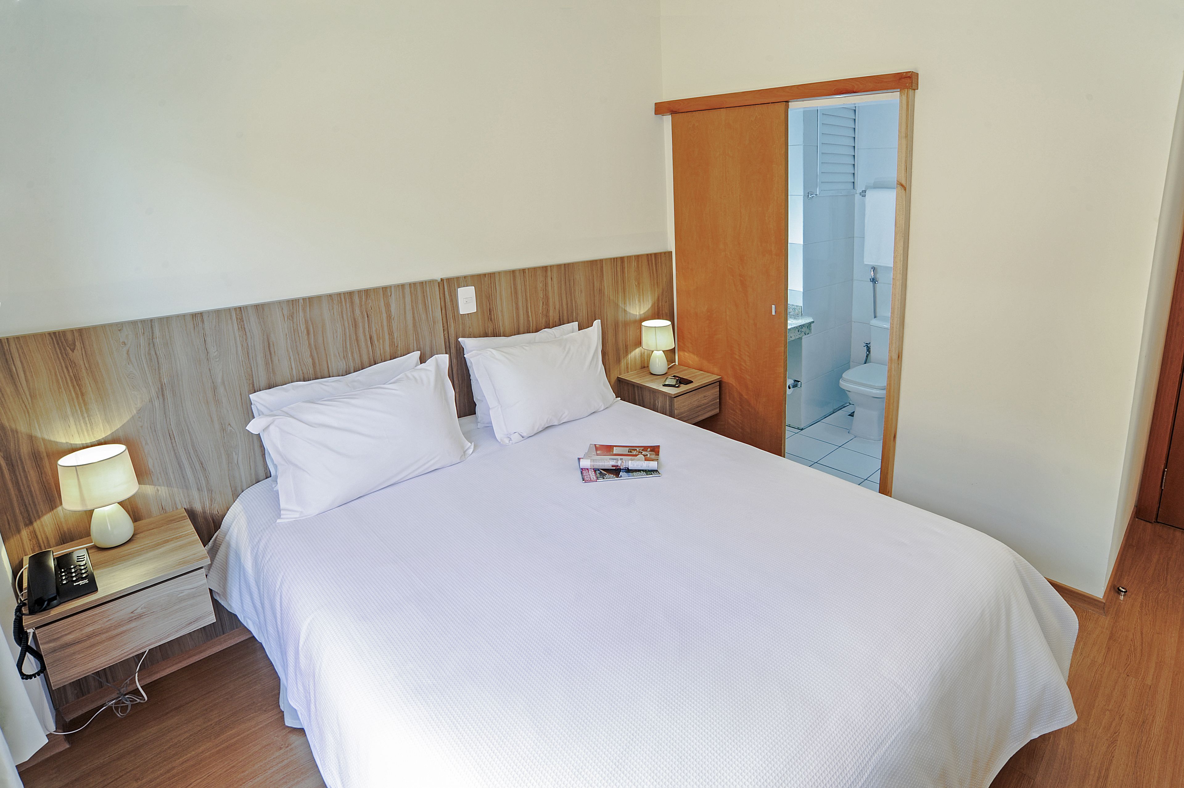 Camera d'albergo con un letto grande, un comodino con una lampada, un telefono e una rivista.