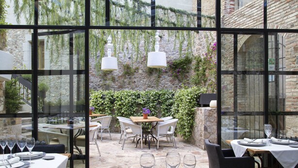 An outdoor dining area with tables and chairs, large windows, and a stone wall with greenery. - Casa Granados Boutique Hotel (Tossa de Mar)