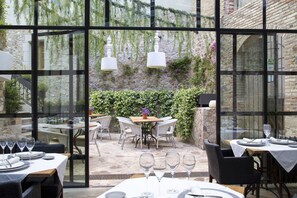 An outdoor dining area with tables and chairs, large windows, and a stone wall with greenery. - Casa Granados Boutique Hotel (Tossa de Mar)