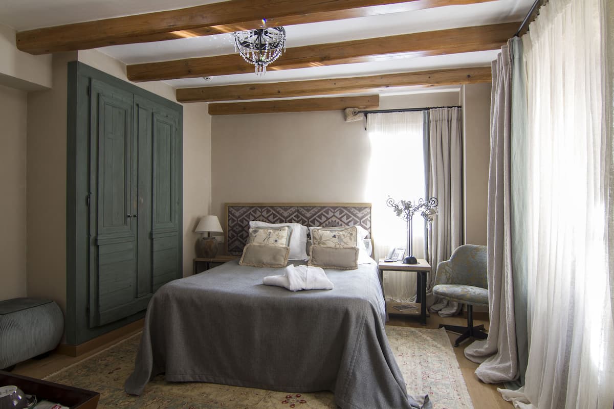 A bedroom with a large bed, a wooden ceiling, a chandelier, a nightstand, a chair, and a wardrobe.