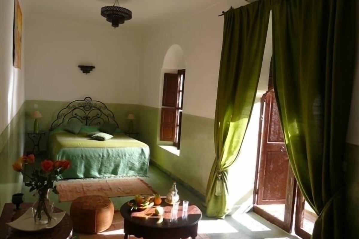 A bedroom with a bed, a nightstand, a table with fruits, a vase with flowers, and a window with curtains.