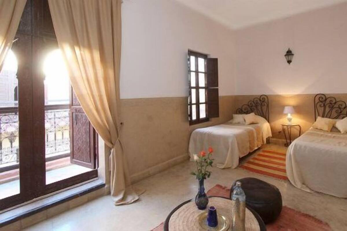 A room with two beds, a window with a view, a small table with a vase, and a lamp.