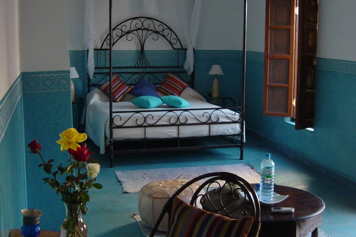 A bedroom with a four-poster bed, a bedside table with a lamp, a chair, and a small table with a vase of flowers.
